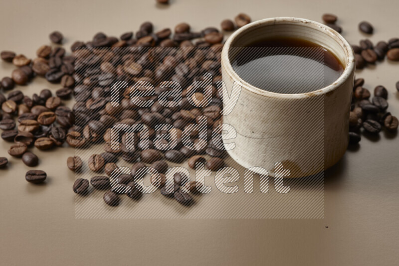 A beige pottery cup of coffee surrounded by roasted coffee beans on beige background
