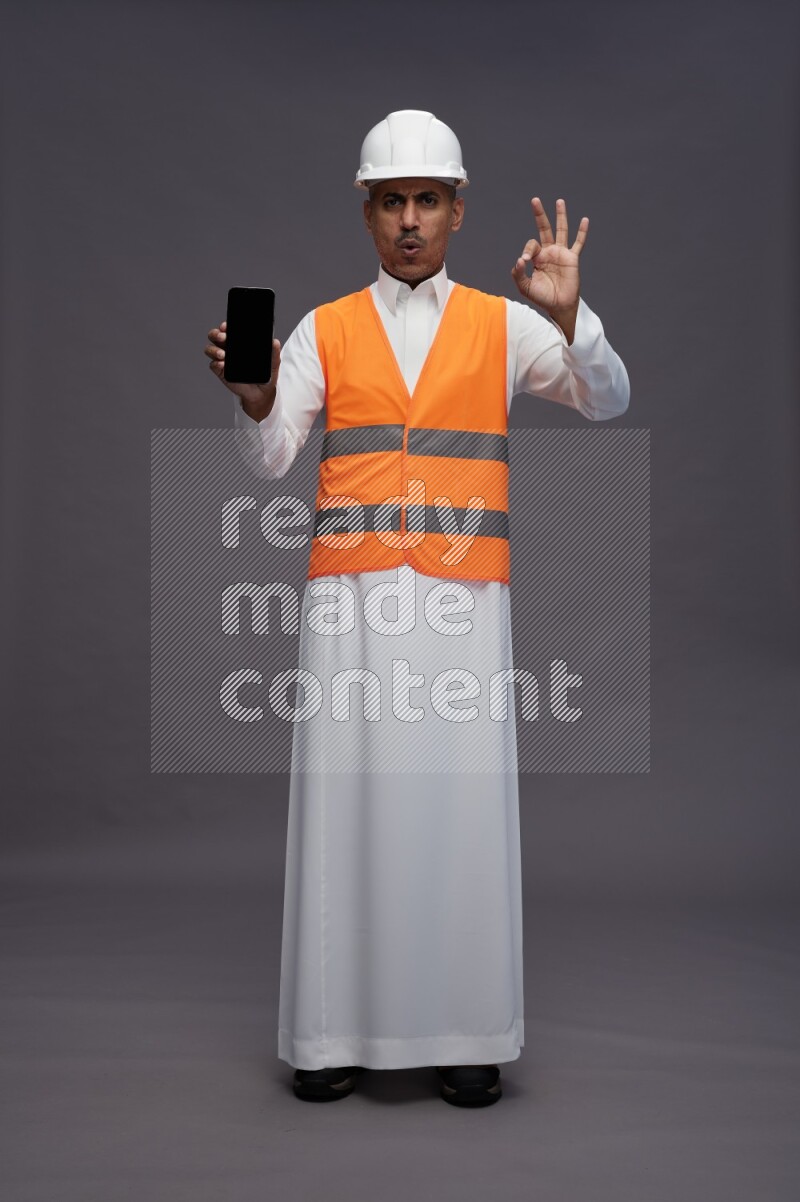 Saudi man wearing thob with engineer vest standing showing phone to camera on gray background