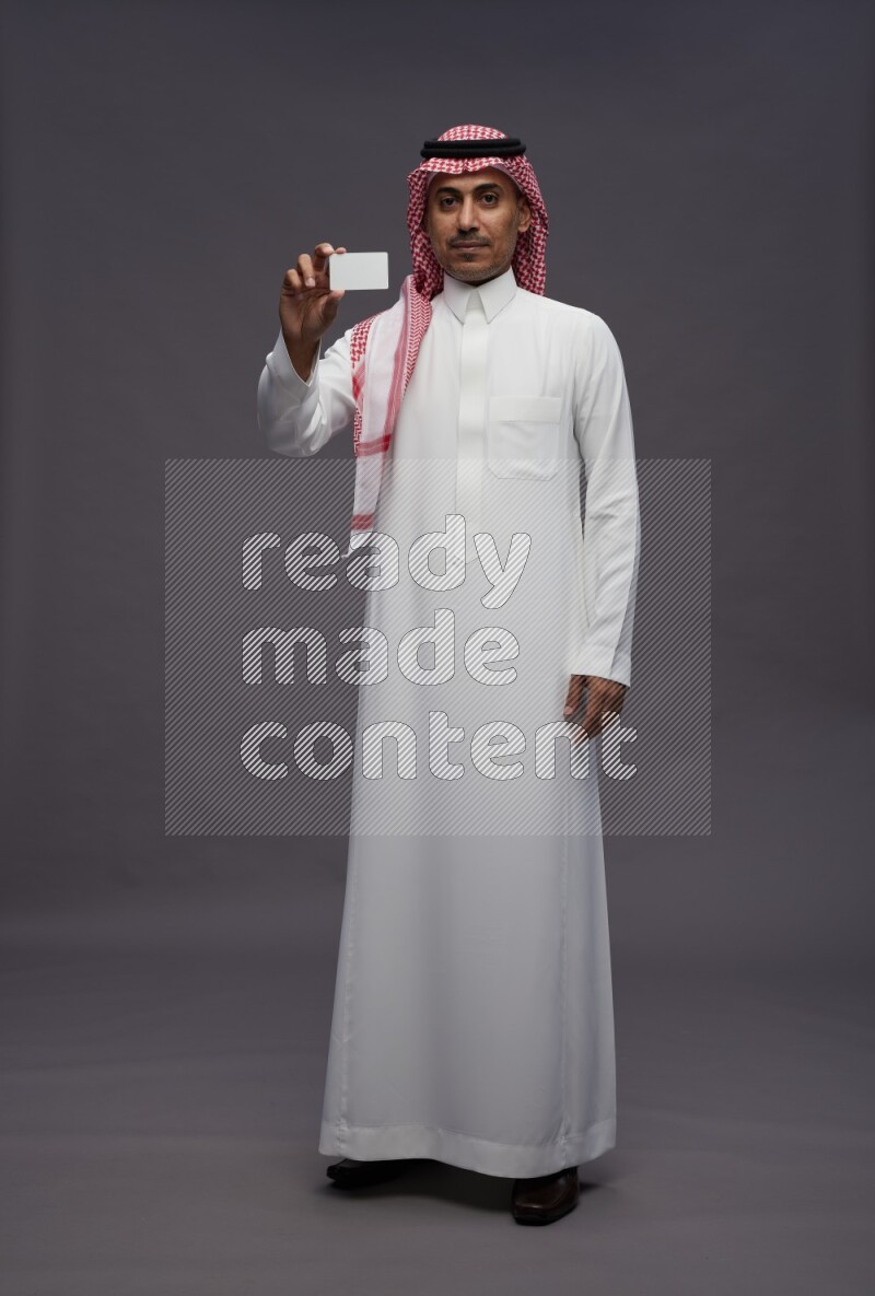 Saudi man wearing thob and shomag standing holding ATM card on gray background