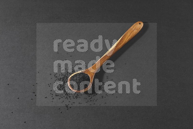 A wooden ladle full of black seeds and the seeds spread beside it on a black flooring