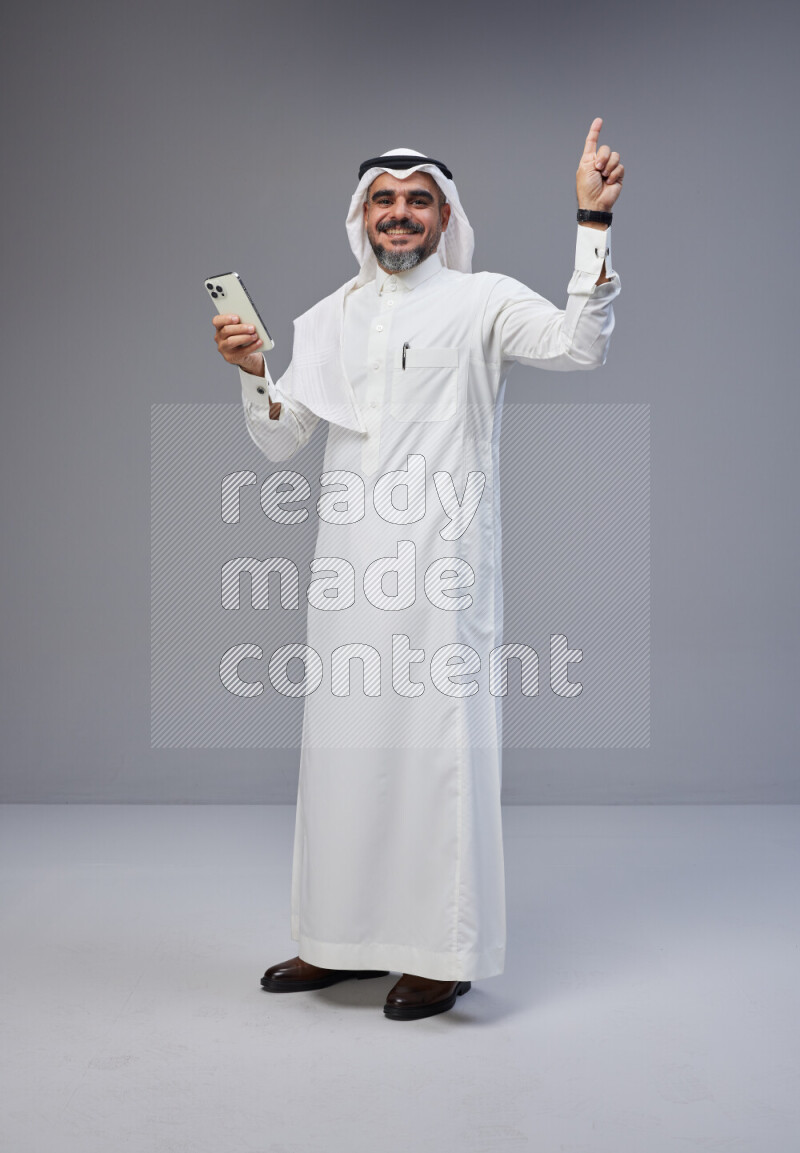 Saudi man Wearing Thob and white Shomag standing texting on phone on Gray background
