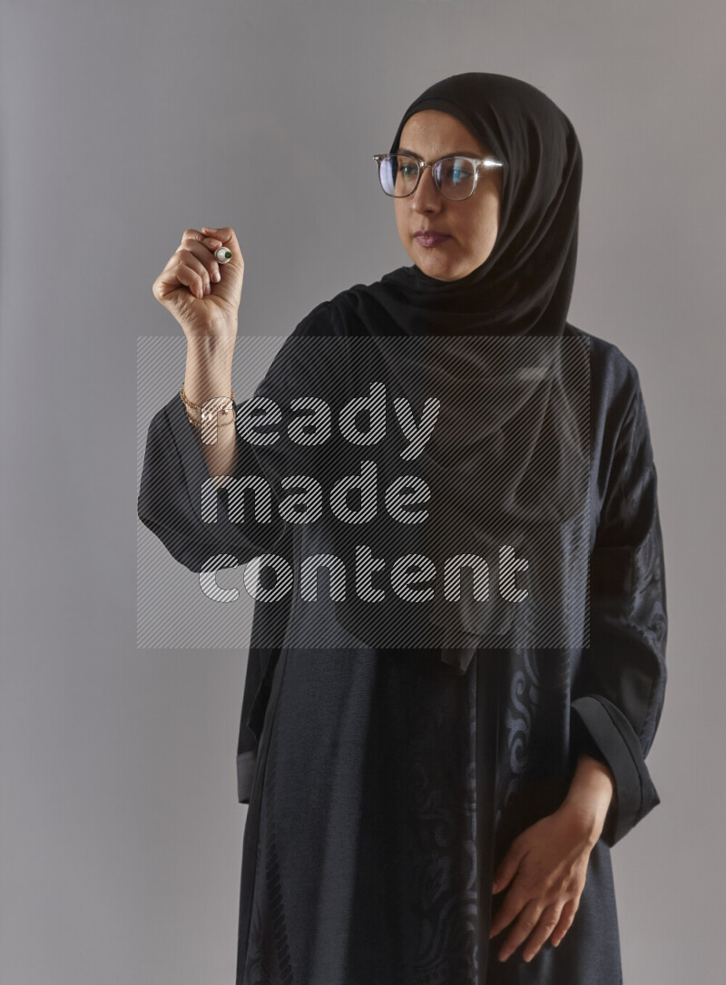 Woman in black abaya, hijab and glasses holding a marker pen to write on a transparent board with different reactions and expressions on grey background