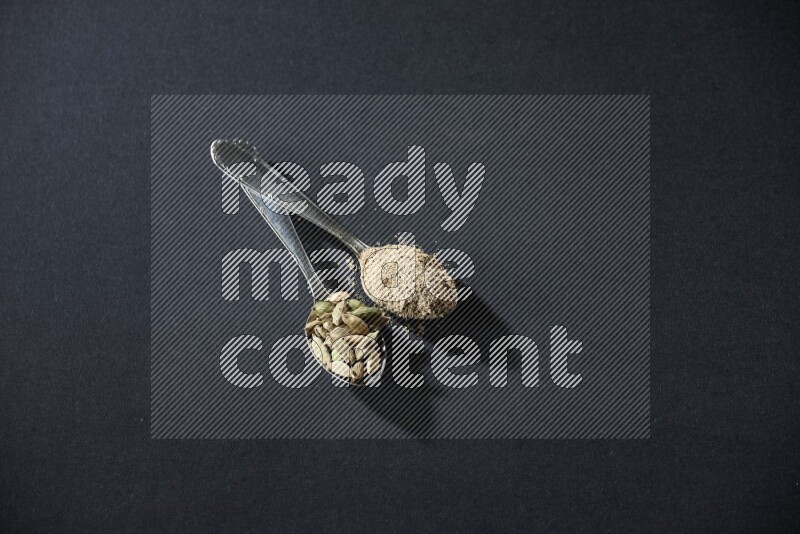 ملعقتان معدنيتان ممتلئتان ببودرة الهيل وحبوب الهيل على خلفية سوداء