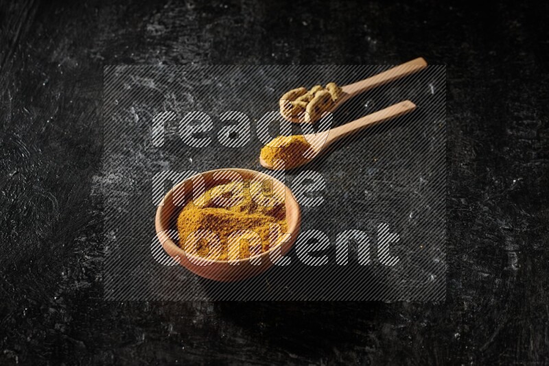 A wooden bowl full of turmeric powder and 2 wooden spoons full of dried turmeric finger and turmeric powder on textured black flooring