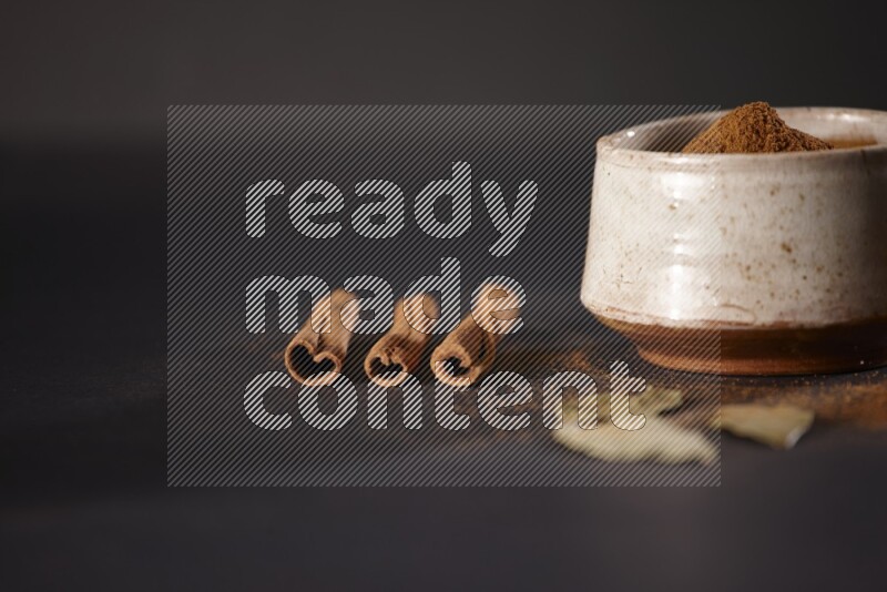 Cinnamon powder in a white pottery bowl and cinnamon sticks and laurel leaves on black background