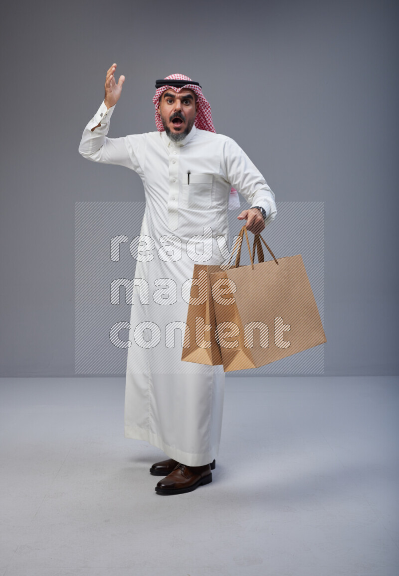 Saudi man Wearing Thob and red Shomag standing holding shopping bag on Gray background