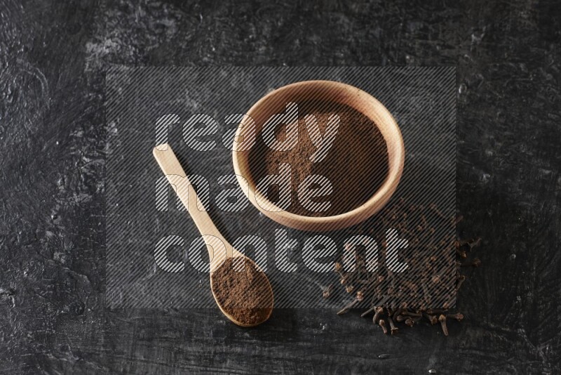 A wooden bowl and a wooden spoon full of cloves powder with spreaded cloves on a textured black flooring