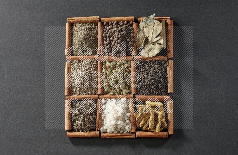 9 مربعات من أعواد القرفة ممتلئة بالهيل في المنتصف محاطة بأصابع الكركم والفلفل الأسود وورق الغار والبهار الحلو والكمون والفلفل الأبيض والريحان المجفف والملح على أرضية سوداء