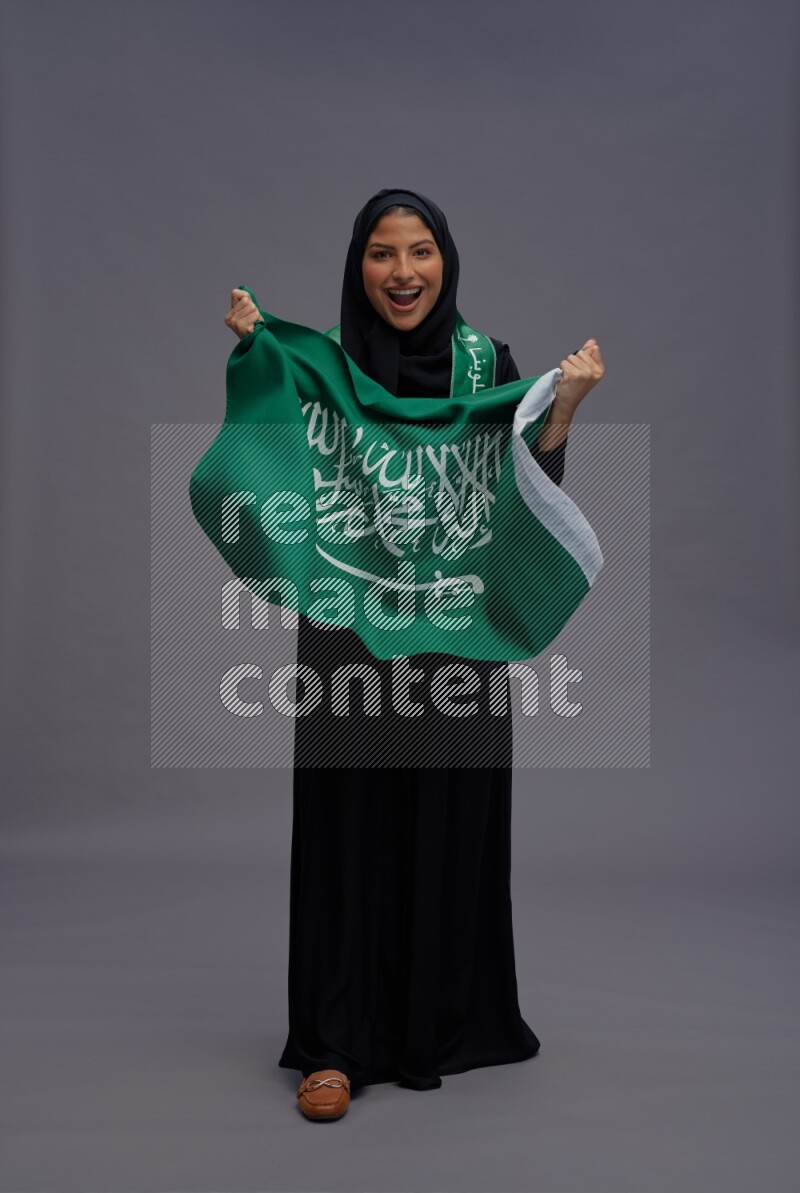 Saudi woman wearing Abaya standing holding Saudi flag on gray background
