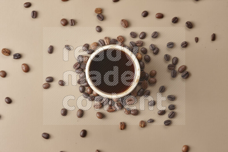 A beige pottery cup of coffee surrounded by roasted coffee beans on beige background