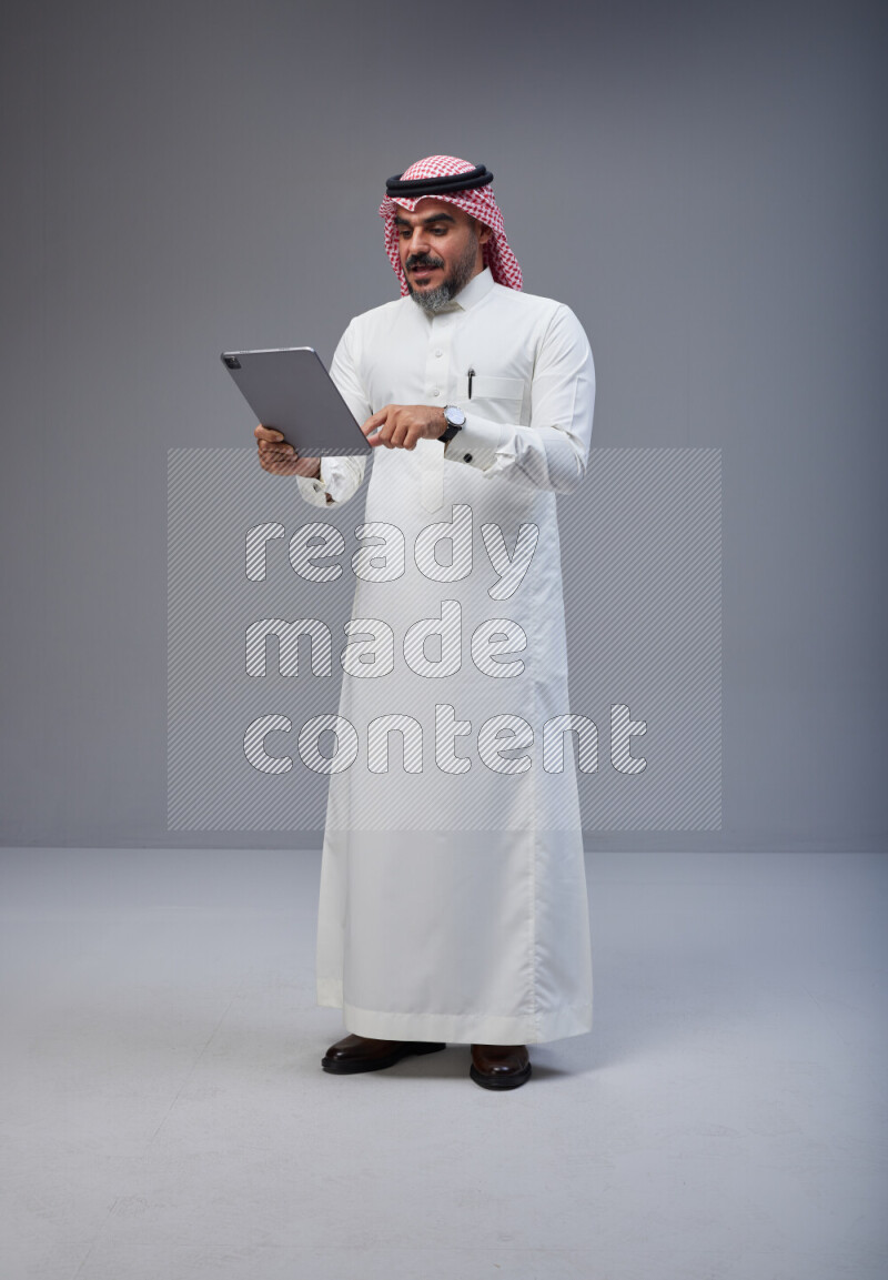 Saudi man Wearing Thob and red Shomag standing working on tablet on Gray background