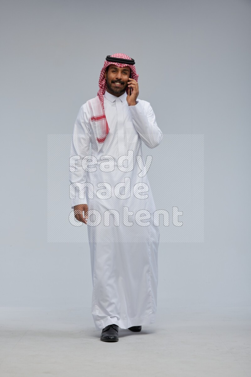 Saudi man Wearing Thob and shomag standing talking on phone on Gray background