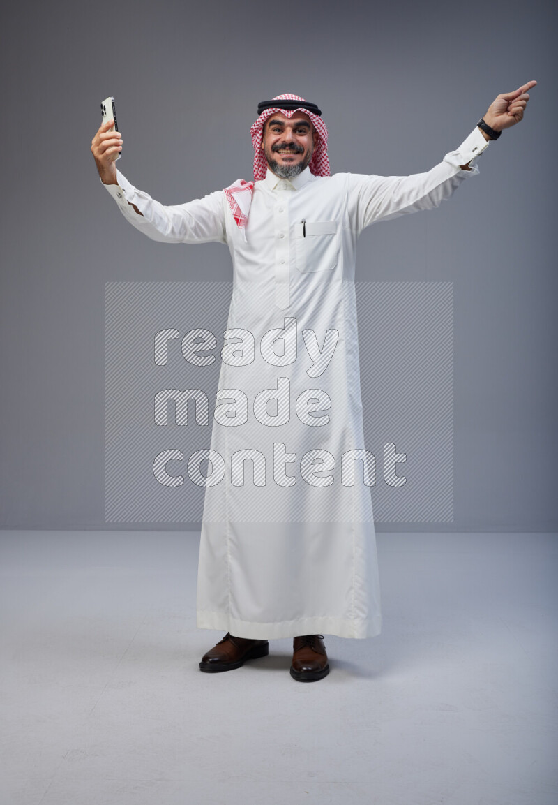 Saudi man Wearing Thob and red Shomag standing taking selfie on Gray background