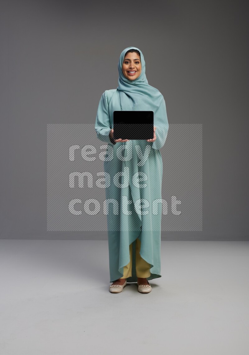 Saudi Woman wearing Abaya standing showing tablet to camera on Gray background