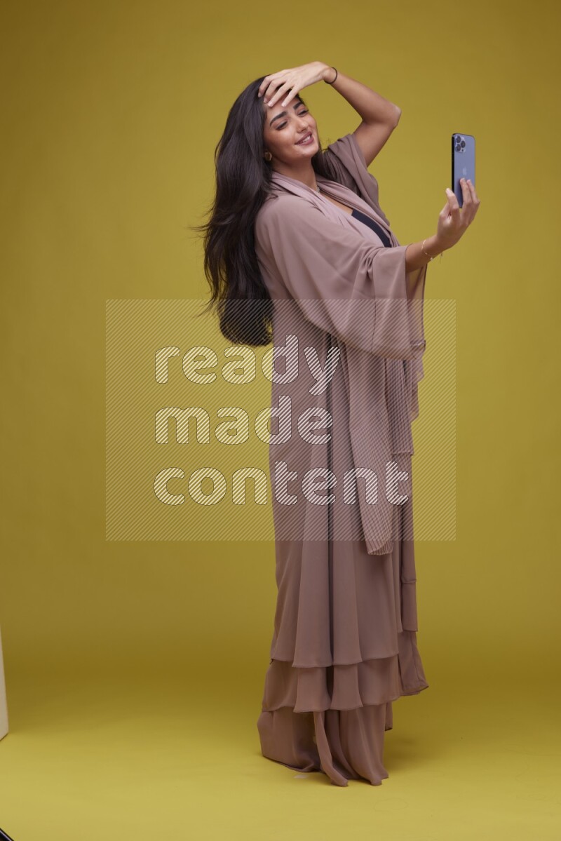 A woman Taking a Selfie on a Yellow Background wearing Brown Abaya