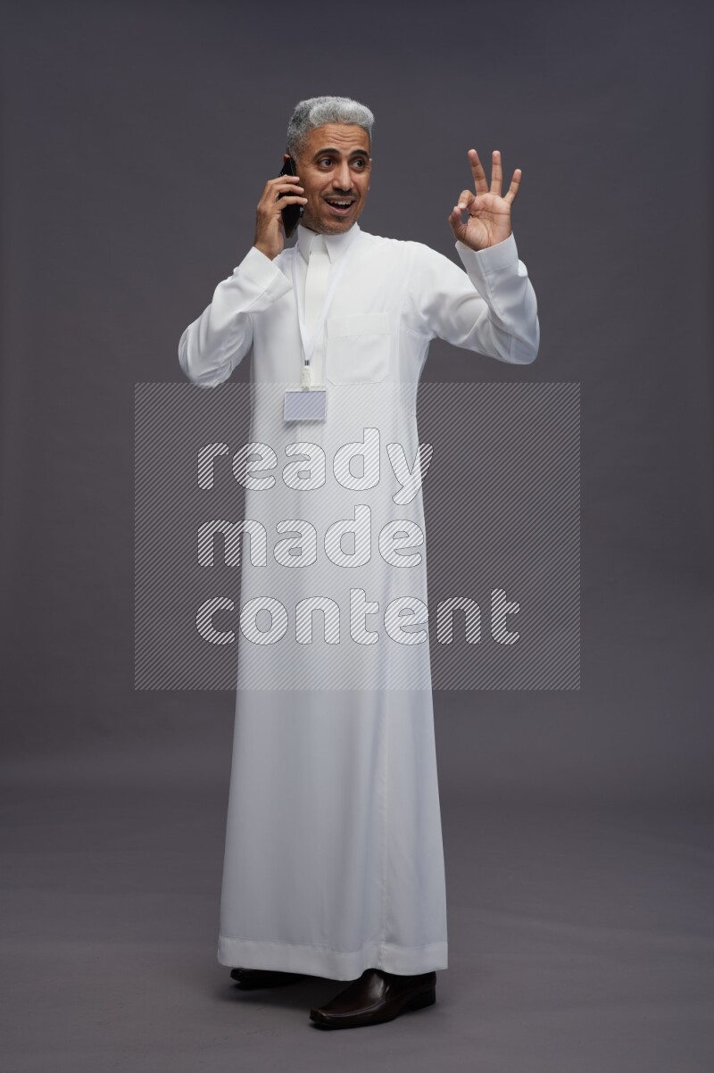 Saudi man wearing thob with neck strap employee badge standing talking on phone on gray background