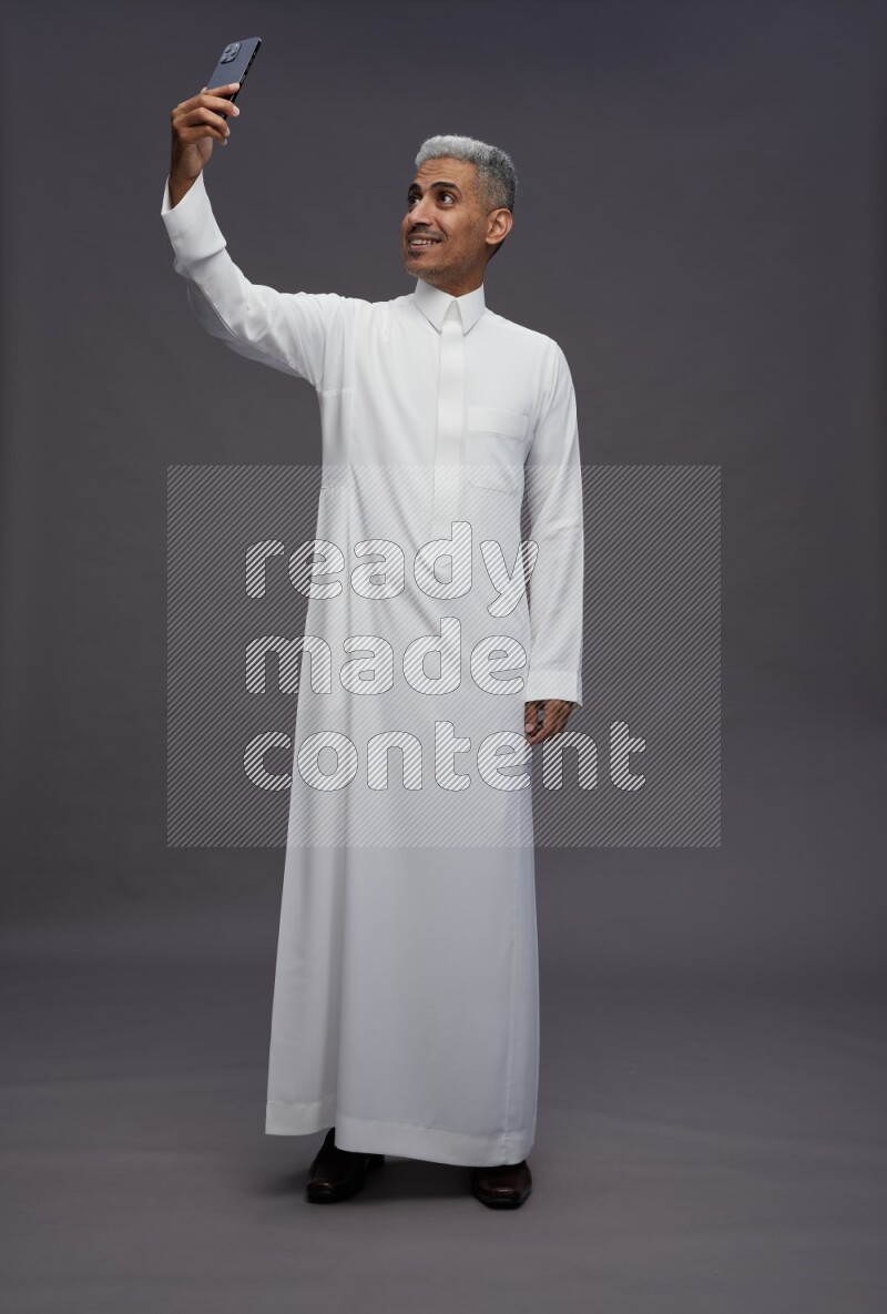 Saudi man wearing thob standing taking selfie on gray background