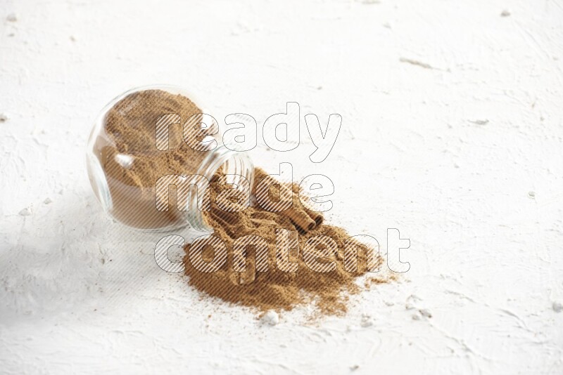 Flipped glass jar full of cinnamon powder with some pieces of cinnamon sticks on a textured white background