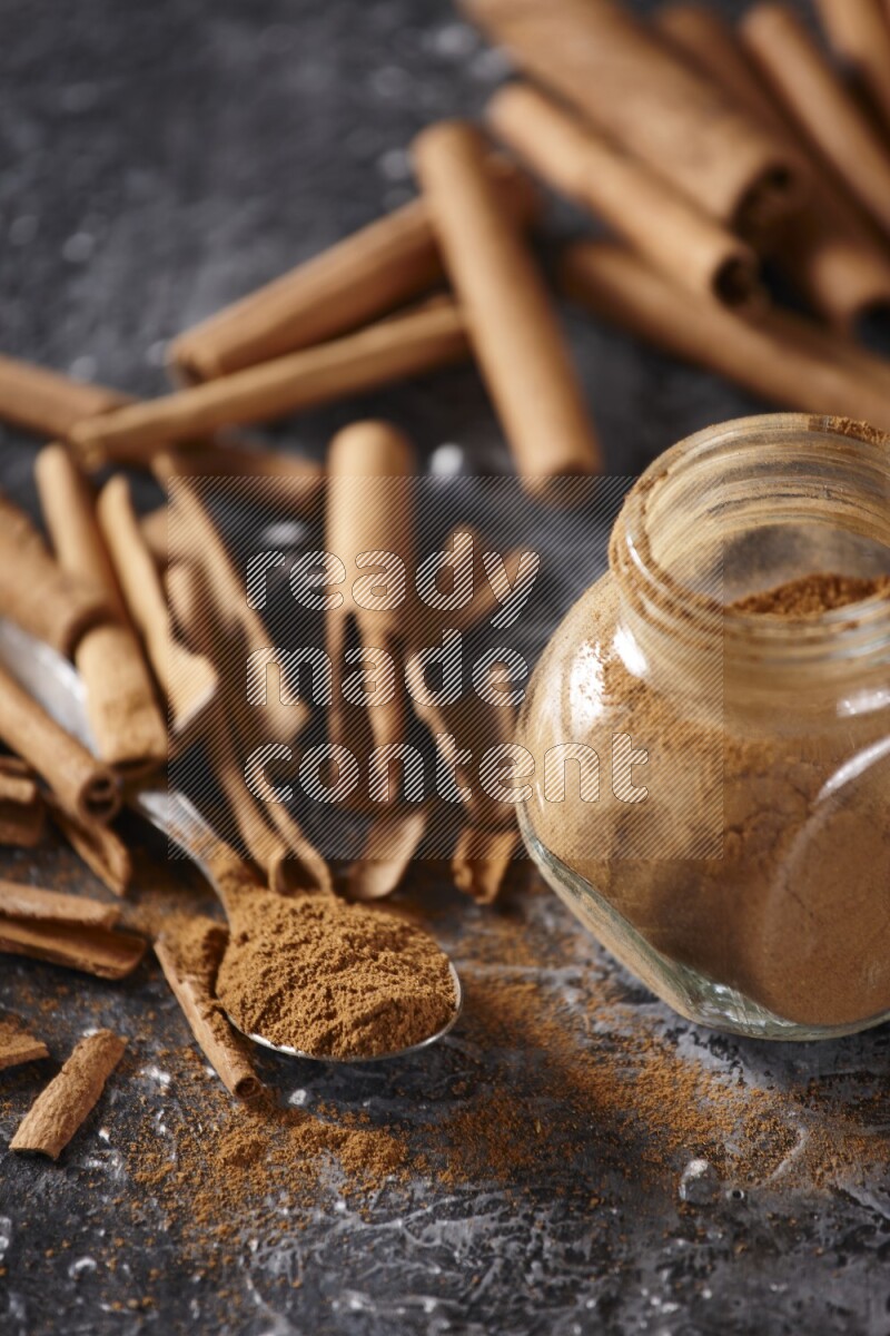 Herbal glass jar and a metal spoon full of cinnamon powder surrounded by cinnamon sticks on textured black background