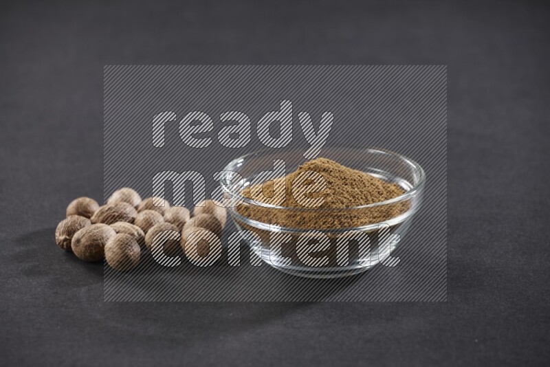 A glass bowl full nutmeg powder with whole seeds beside it on a black flooring