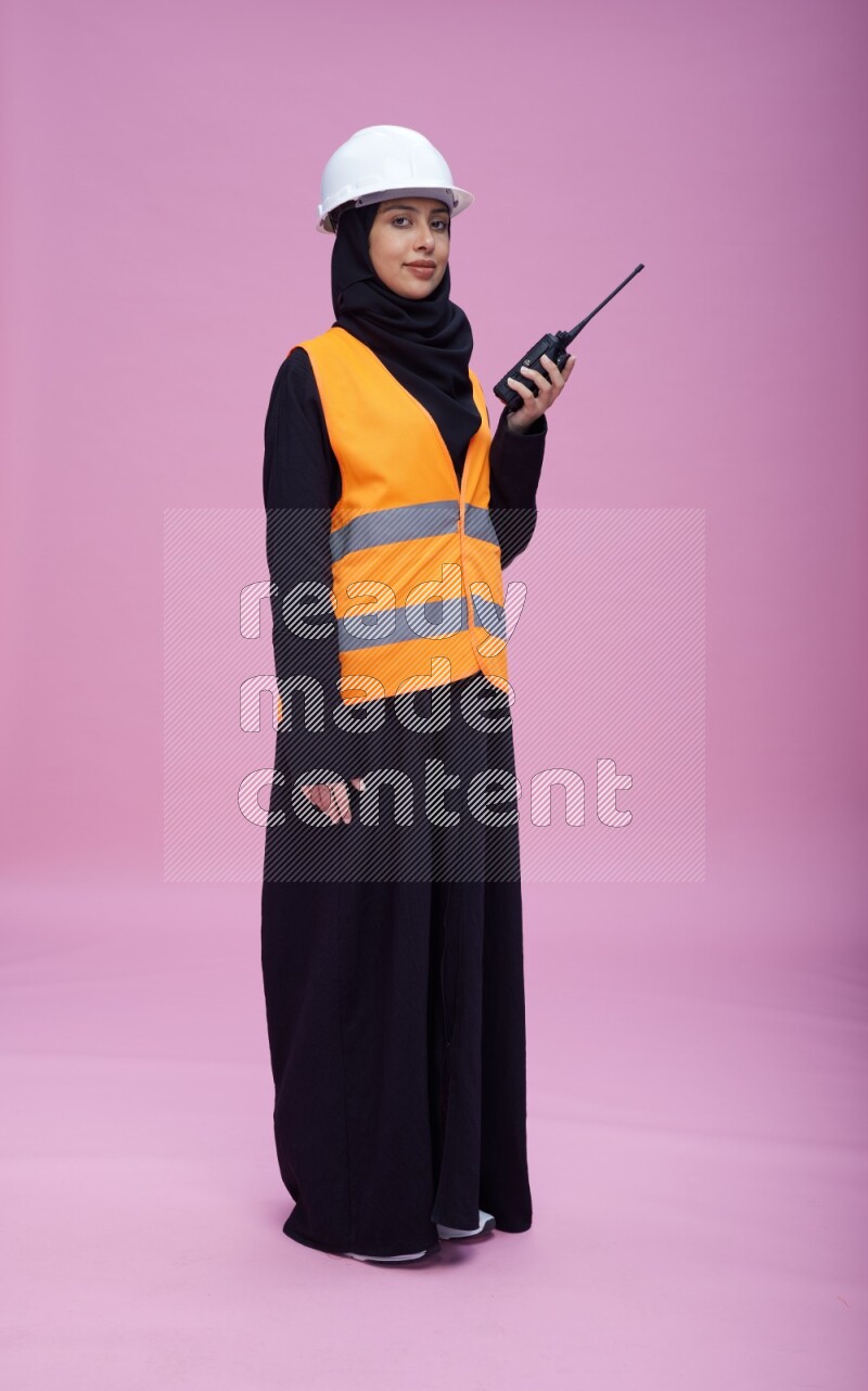 Saudi woman wearing Abaya with engineer vest and helmet standing holding walkie-talkie on pink background