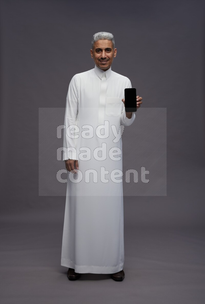 Saudi man wearing thob standing showing phone to camera on gray background