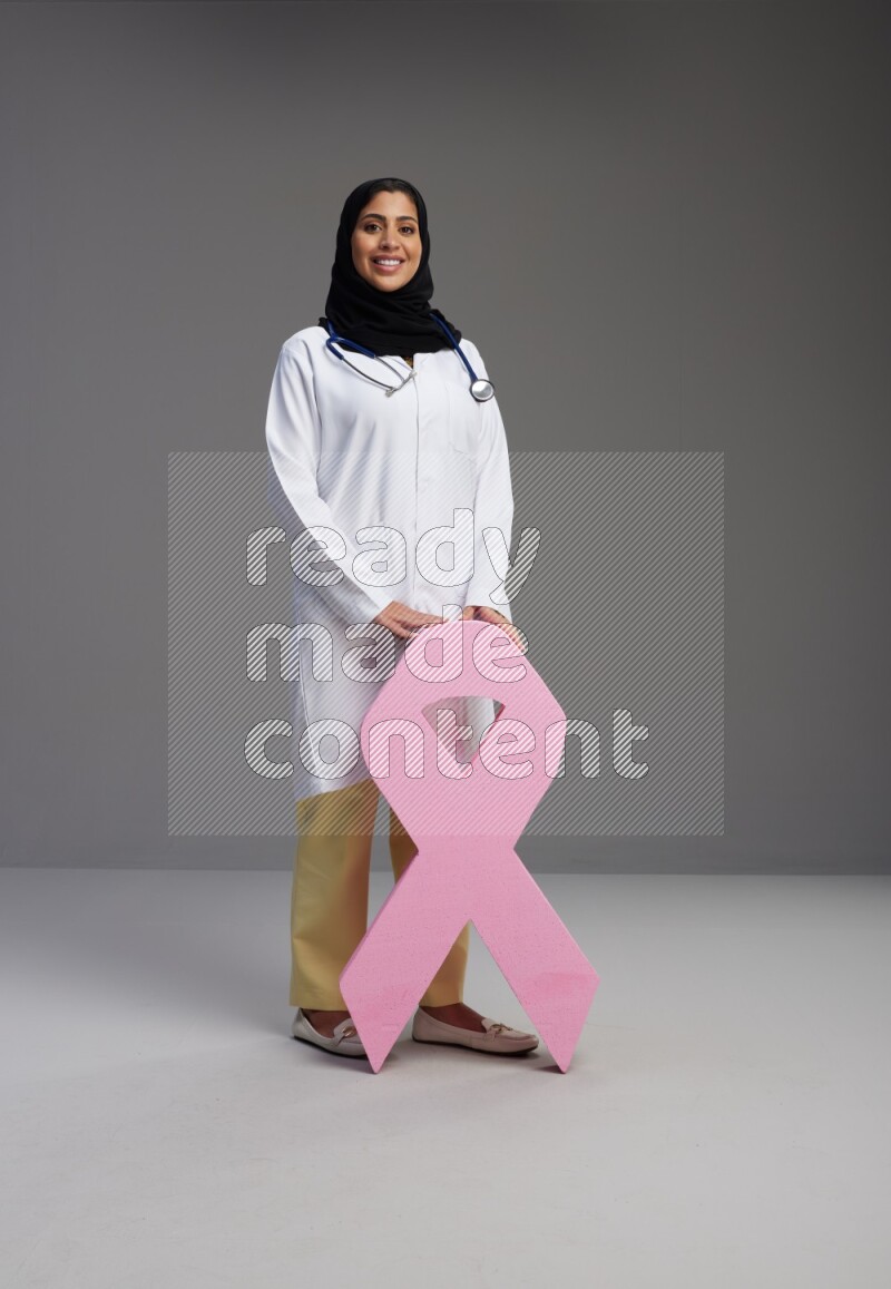 Saudi woman wearing lab coat with stethoscope standing holding awareness ribbon symbol standing on Gray background