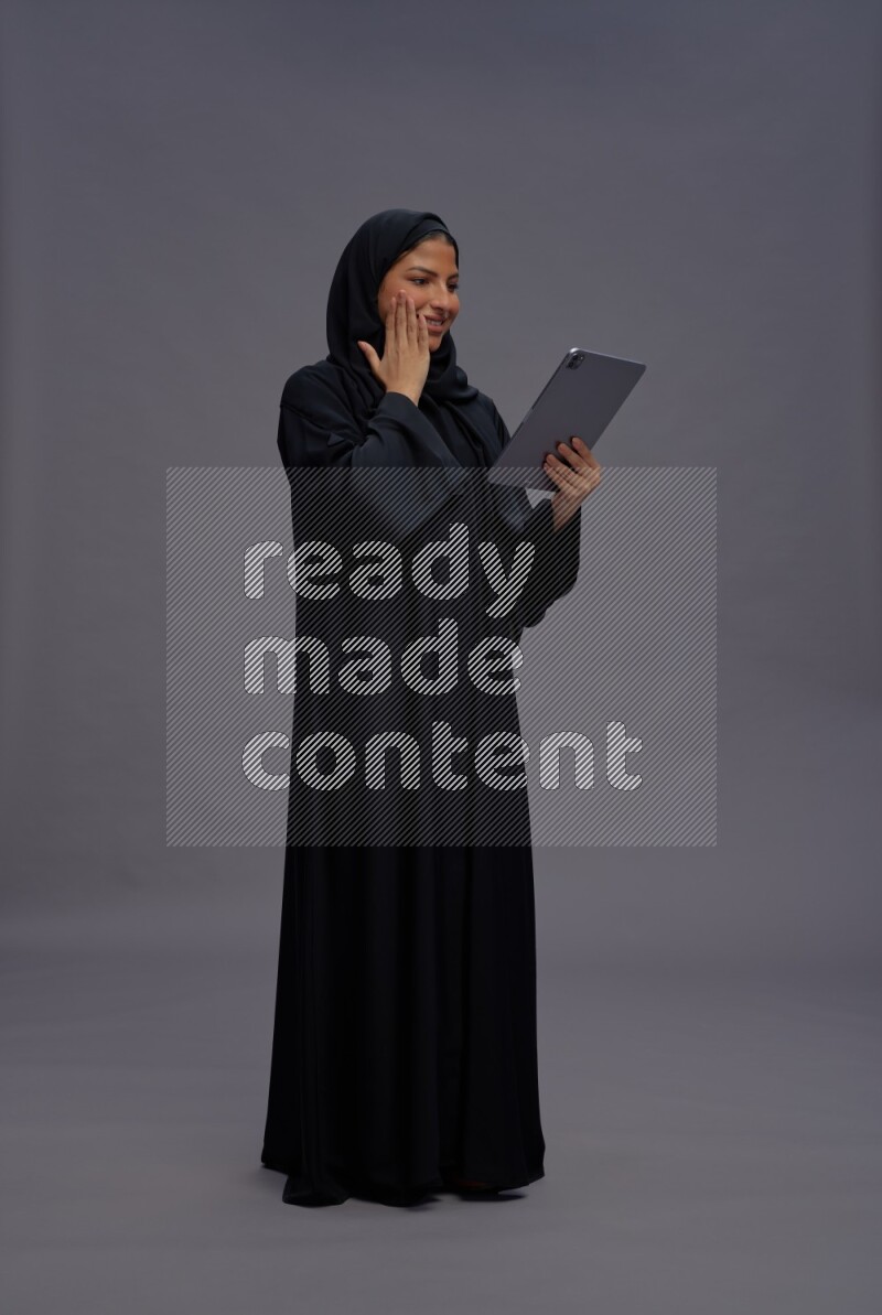 Saudi woman wearing Abaya standing working on tablet on gray background