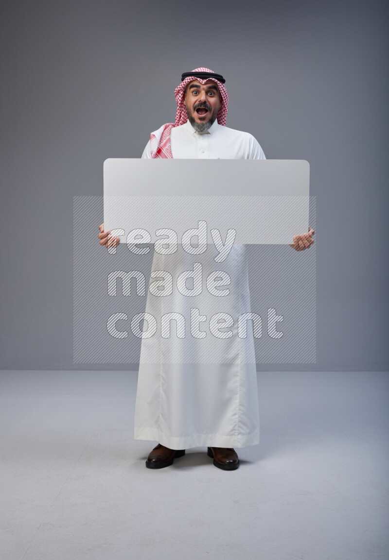 Saudi man Wearing Thob and red Shomag standing holding board on Gray background