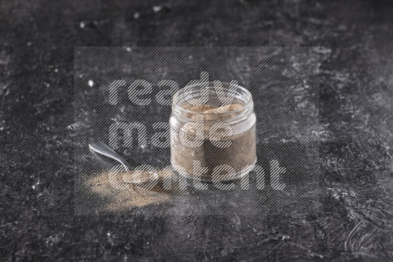 A glass jar full of black pepper powder and a metal spoon full of powder on a textured black flooring