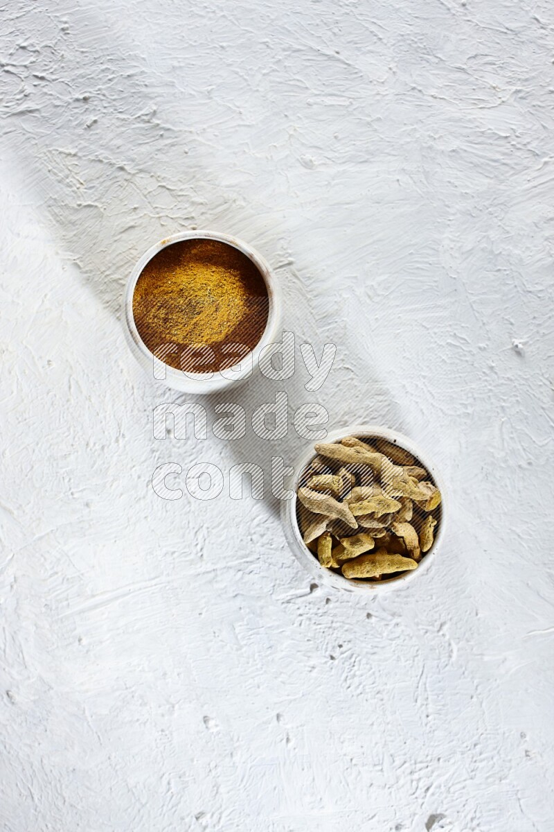 وعاءان فخاريان بيج ممتلئان ببودرة الكركم وأصابع الكركم المجففة على خلفية بيضاء