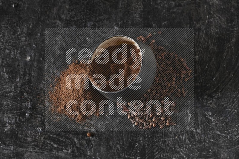 A black pottery bowl full of cloves powder spilled out of it with whole grains on a textured black flooring