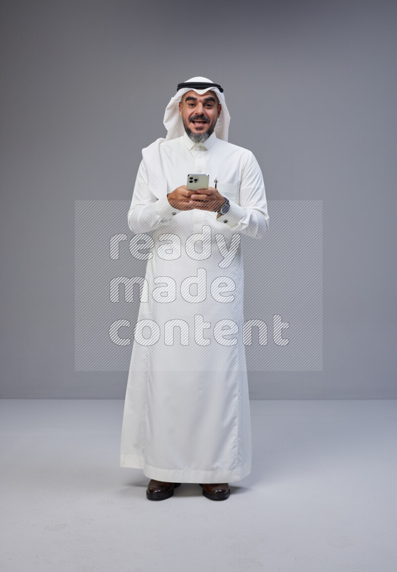 Saudi man Wearing Thob and white Shomag standing texting on phone on Gray background
