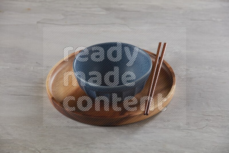Blue pottery round bowl on top of brown wood round plate and wood chopsticks, on grey textured countertop
