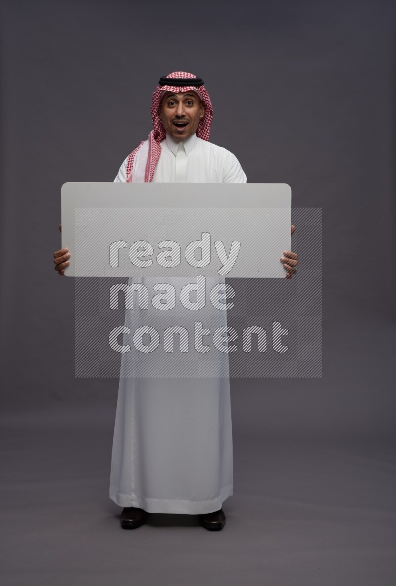 Saudi man wearing thob and shomag standing holding board on gray background