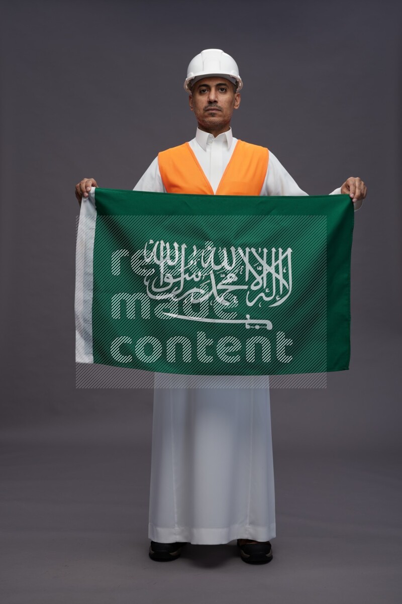 Saudi man wearing thob with engineer vest standing holding Saudi flag on gray background