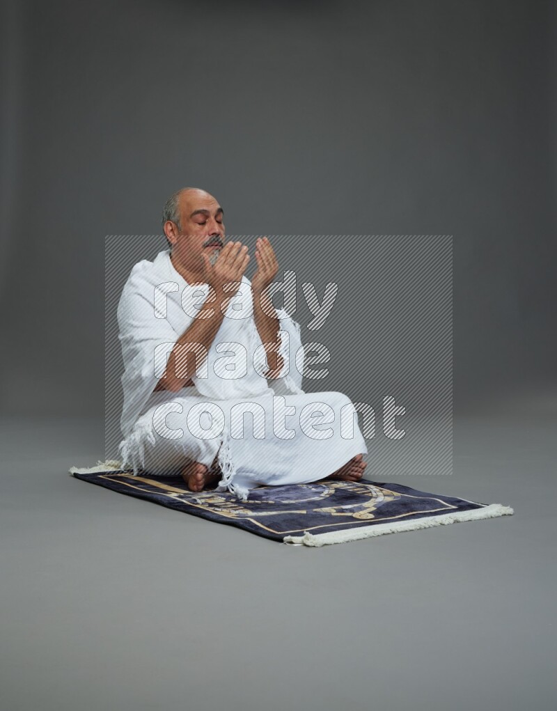 A man wearing Ehram sitting on prayer mat dua'a on gray background