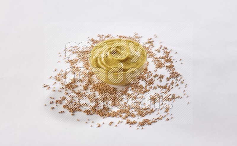 وعاء زجاجي ممتلئ بمعجون الخردل مع بعض بذور الخردل تحت الوعاء على أرضية بيضاء