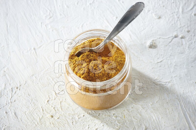 A glass jar and a metal spoon full of turmeric powder on a textured white flooring