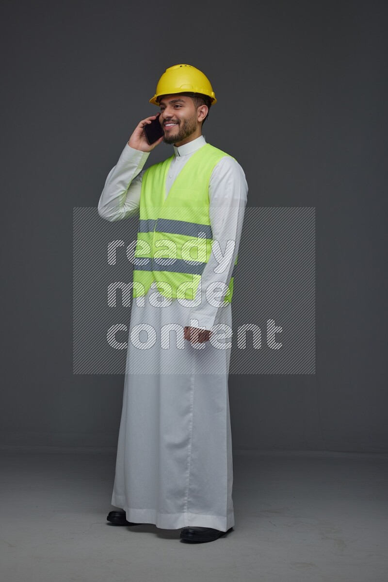 A Saudi man wearing Thobe with a yellow safety vest and white helmet standing and talking in the phone eye level on a gray background