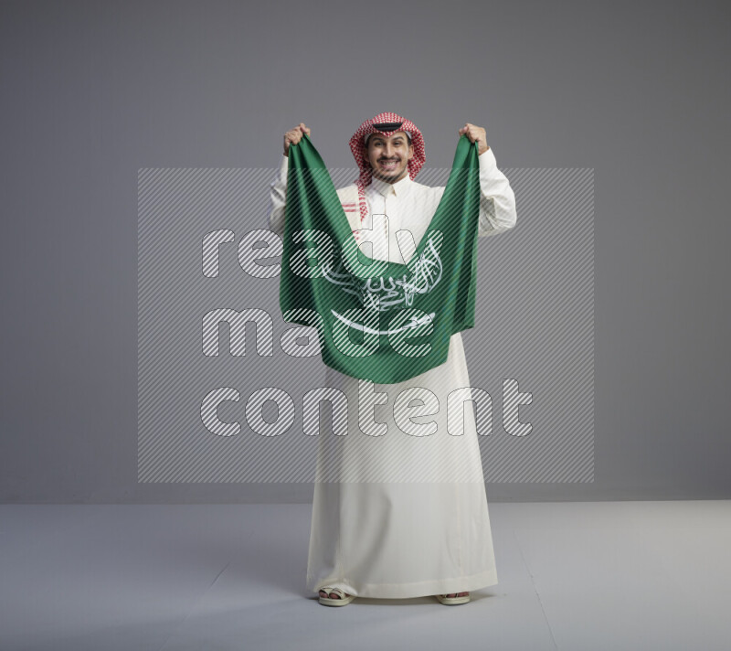A saudi man standing wearing thob and red shomag holding big saudi flag on gray background