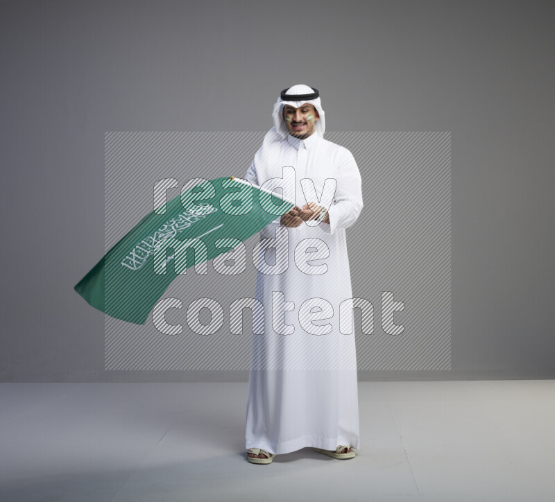 A Saudi man standing wearing thob and white shomag with face painting raising big Saudi flag on gray background