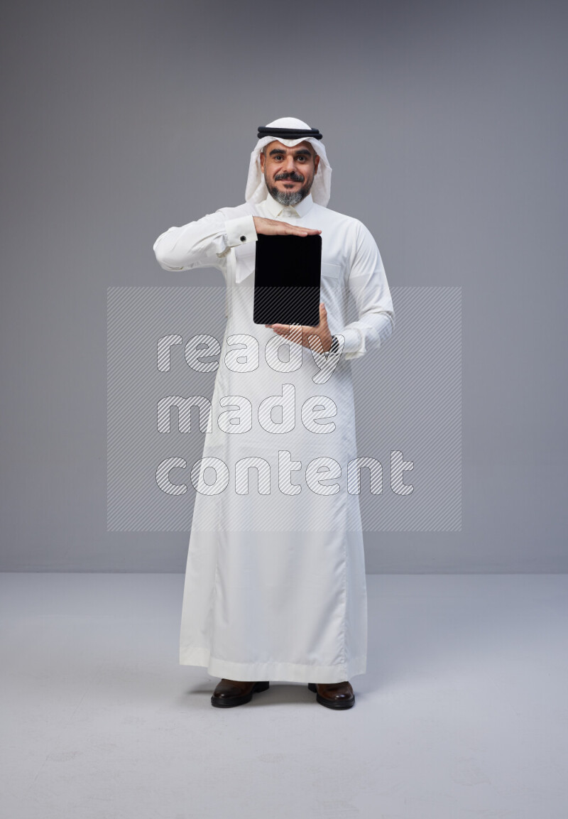 Saudi man Wearing Thob and white Shomag standing showing tablet to camera on Gray background