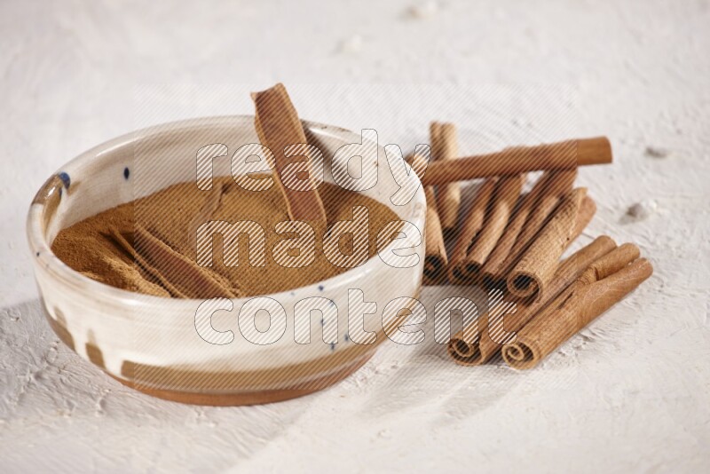 Ceramic bowl full of cinnamon powder with cinnamon sticks on the side on white background
