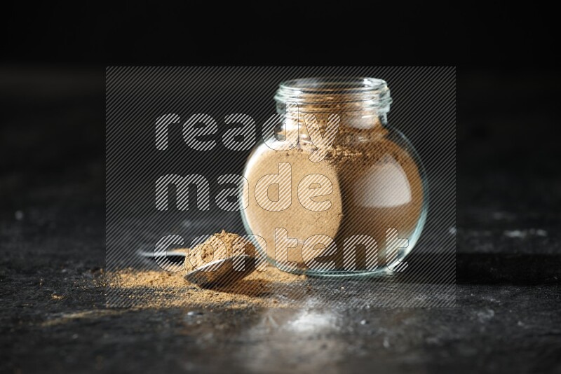 A glass spice jar and metal spoon full of allspice powder on a textured black flooring