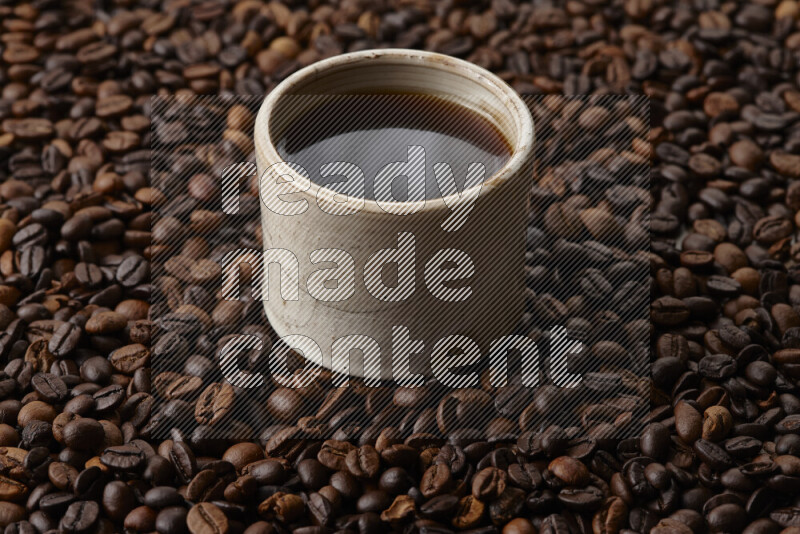 A beige pottery cup of coffee surrounded by roasted coffee beans on beige background