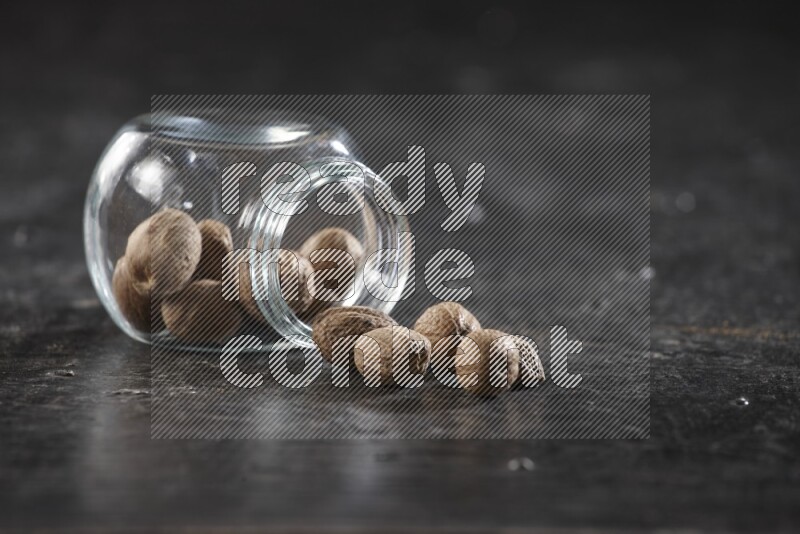 وعاء زجاجي ممتلئ بجوزة الطيب مقلوب وبعض الحبوب متناثرة على خلفية سوداء