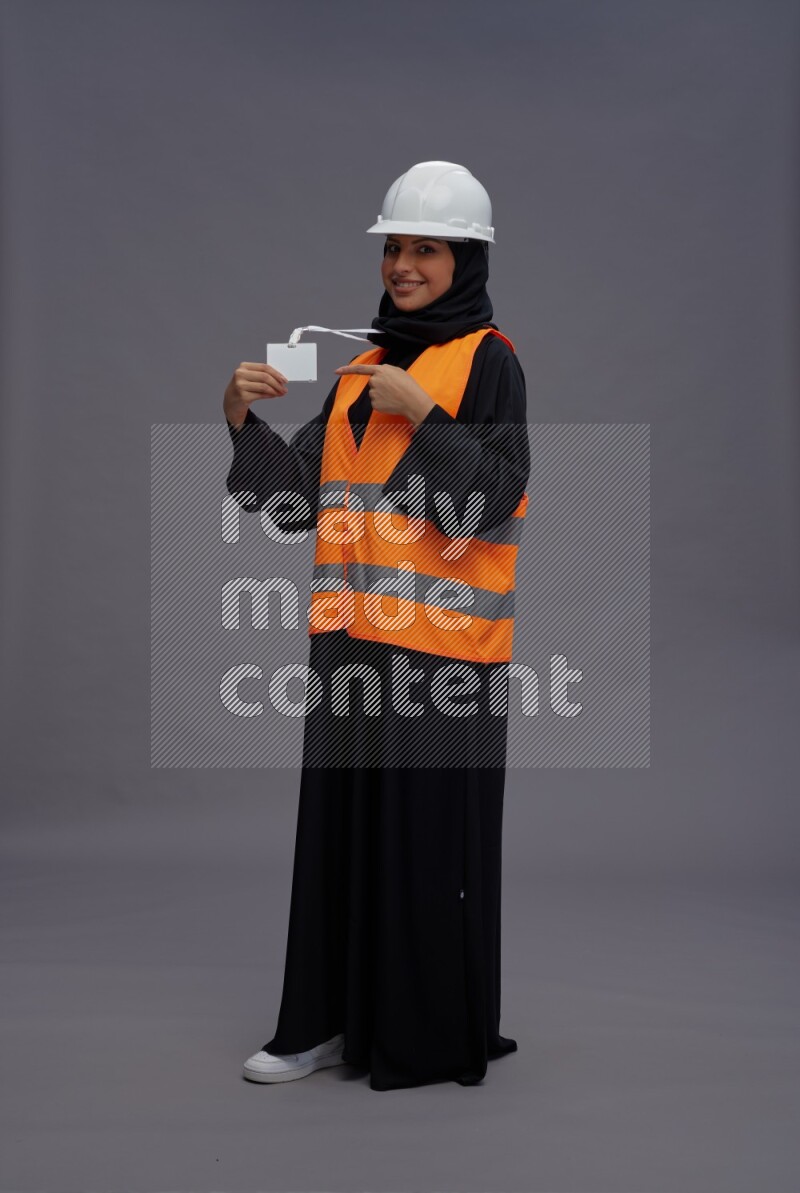 Saudi woman wearing Abaya with engineer vest with neck strap employee badge standing interacting with the camera on gray background