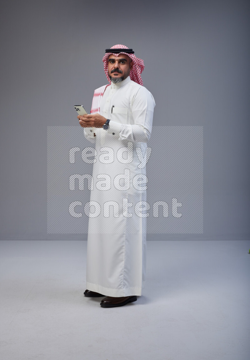 Saudi man Wearing Thob and red Shomag standing texting on phone on Gray background