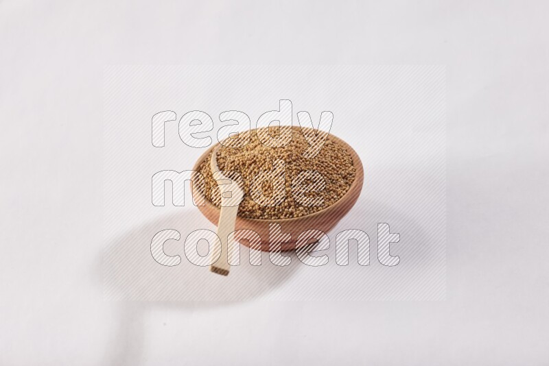 A wooden bowl full of mustard seeds with a wooden spoon on the seed on a white flooring
